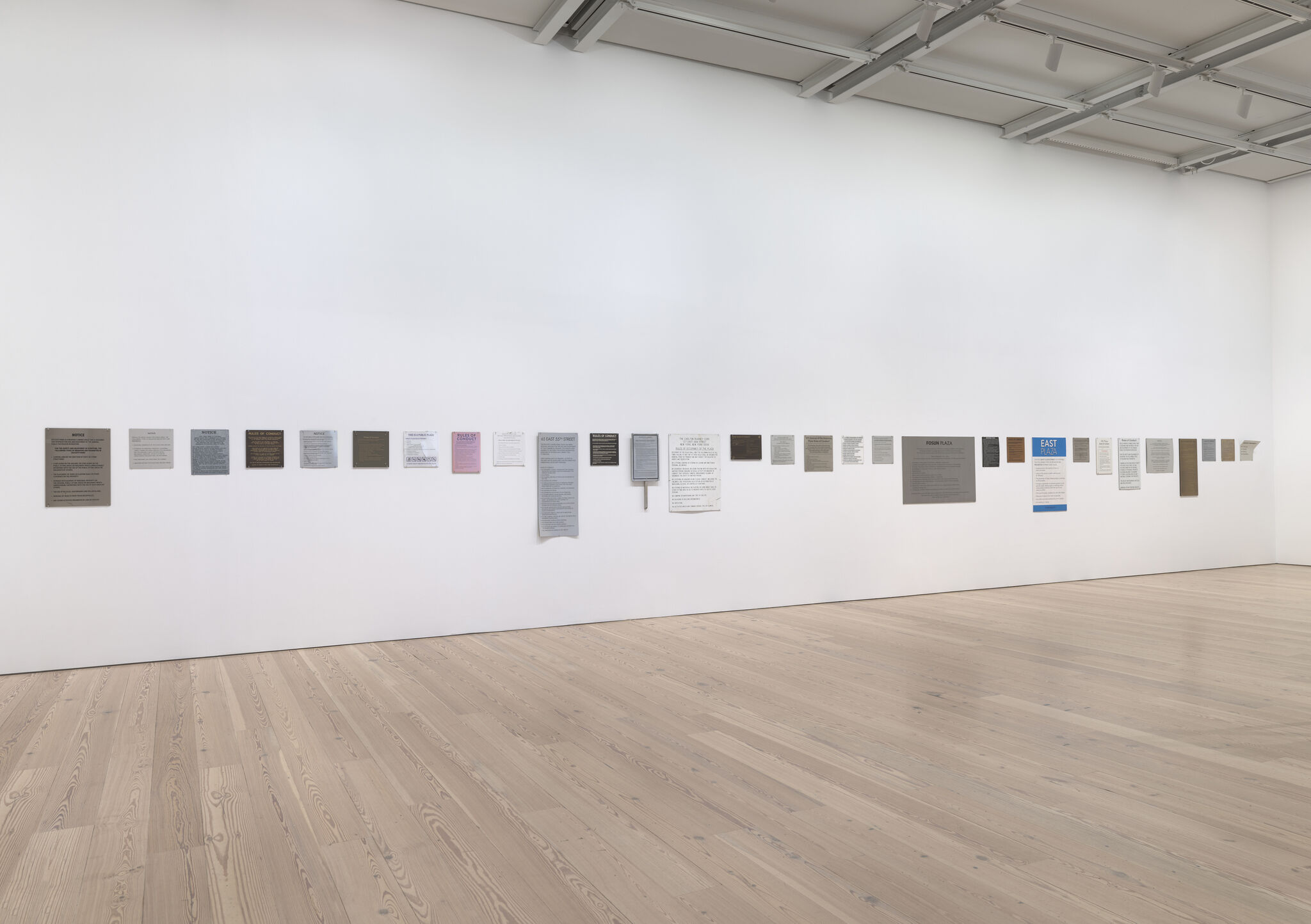 A long row of small plaques and notices displayed low along a large white gallery wall.
