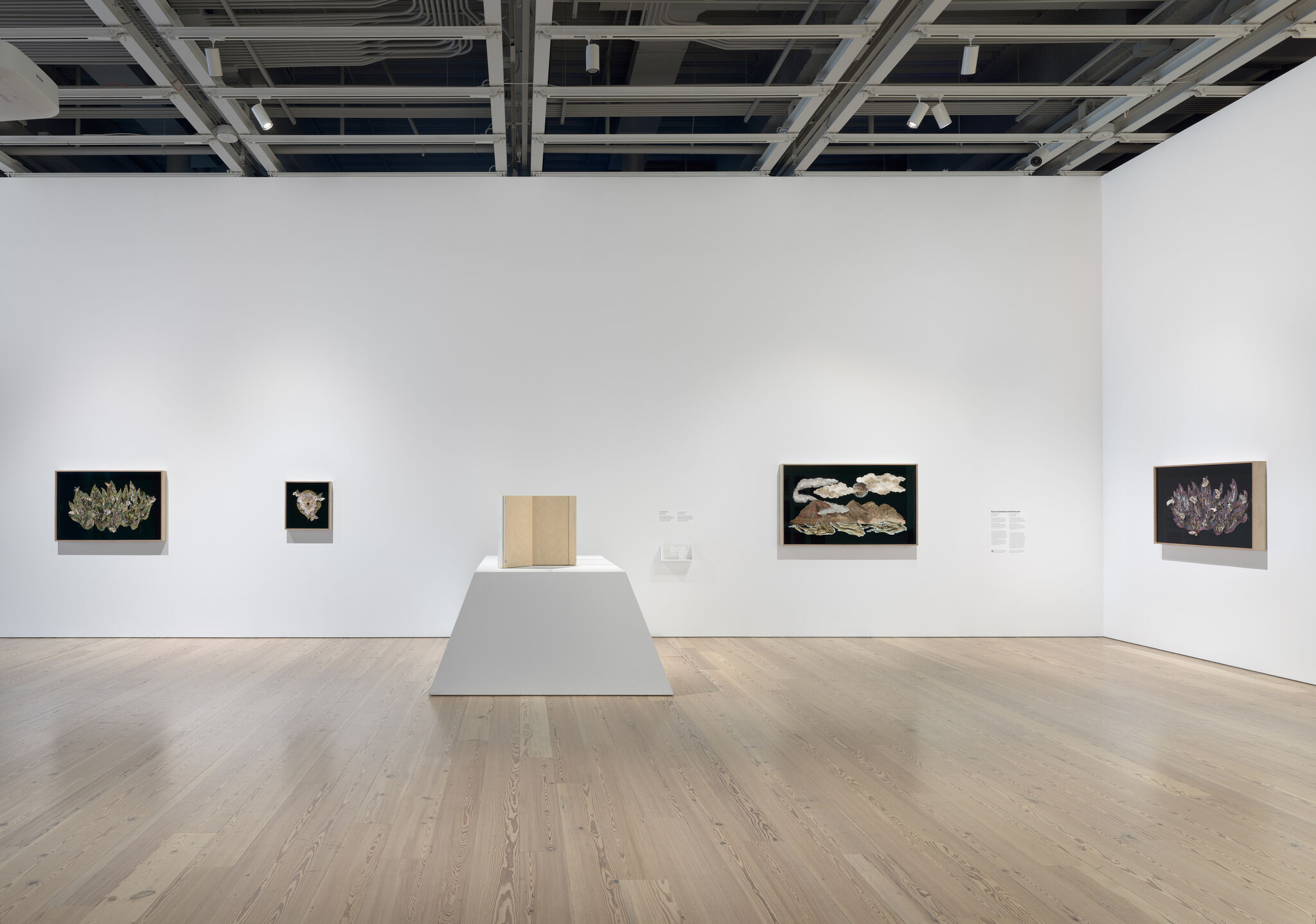 White gallery wall displaying four framed artworks and a wooden box on a central pedestal.