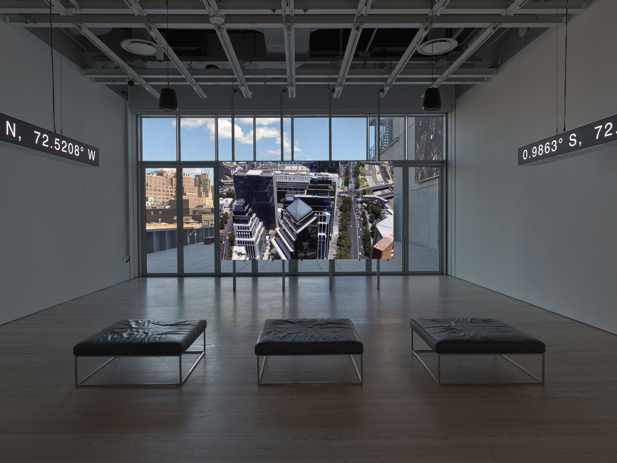 Three benches face a large screen showing an aerial city view in a dim gallery.