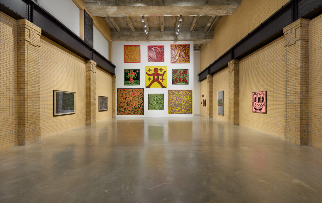Installation view of Keith Haring at The Brant Foundation New York, a big room with 9 paintings on the white wall directly across from the camera and on the side is yellow brick walls with Haring paintings as well.