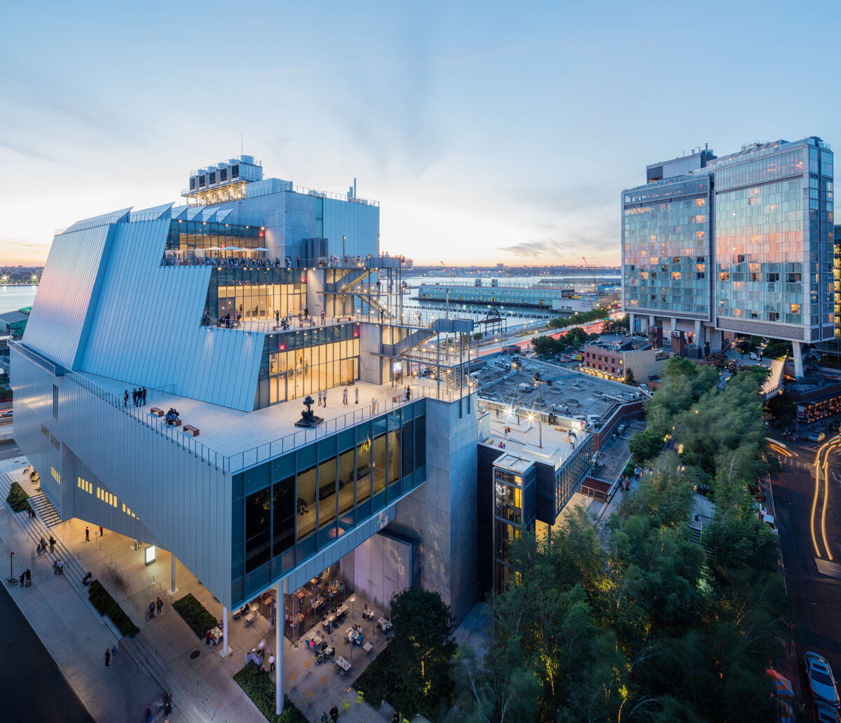 The modern Whitney Museum building that looks very modern with a lot of windows and outdoor terraces and people overlooking the waterfront at dusk.