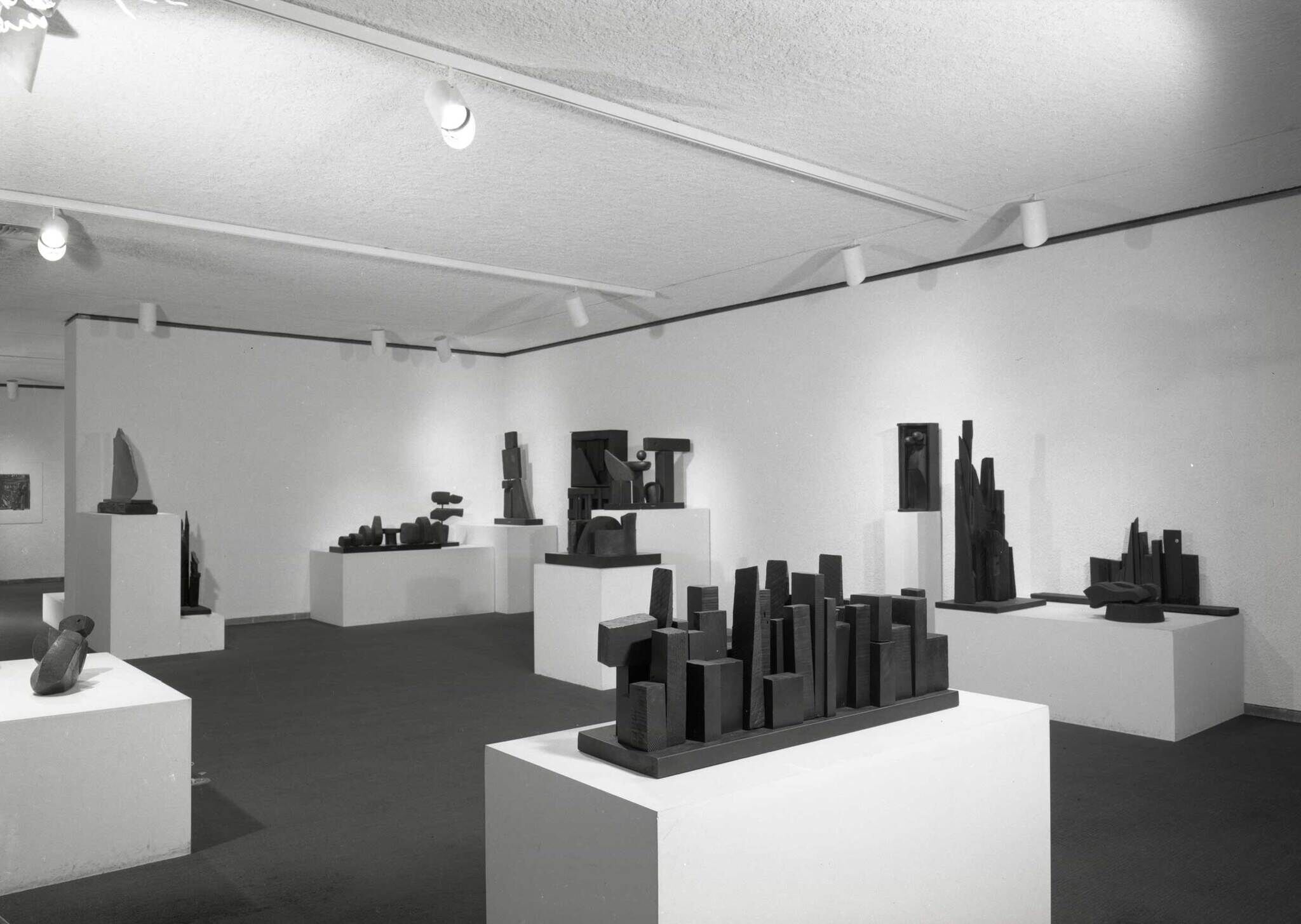 Gallery room with many abstract wooden sculptures displayed on white pedestals under track lighting.