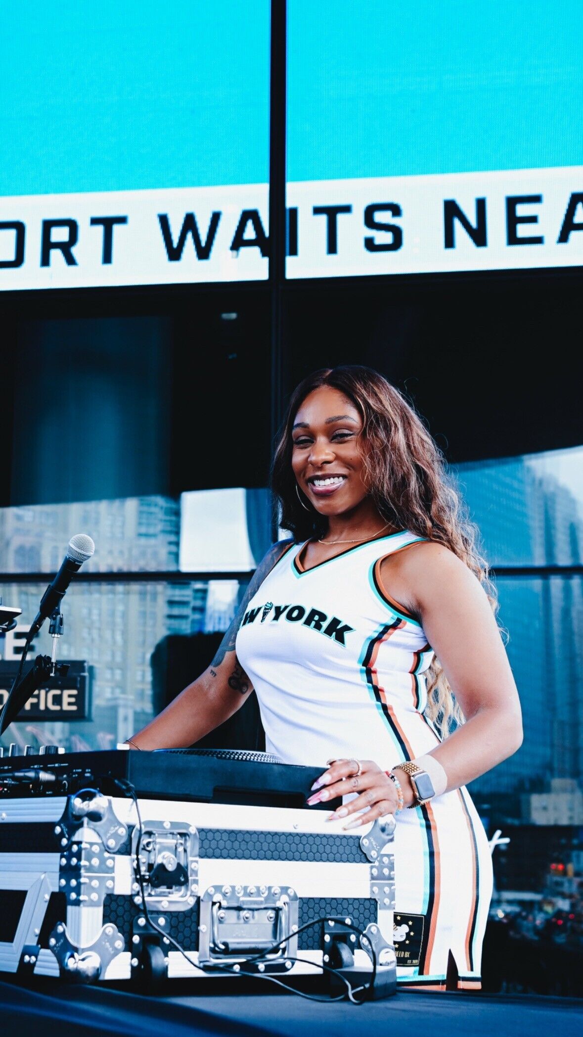 A smiling DJ wearing a New York jersey stands behind turntables and DJ equipment.