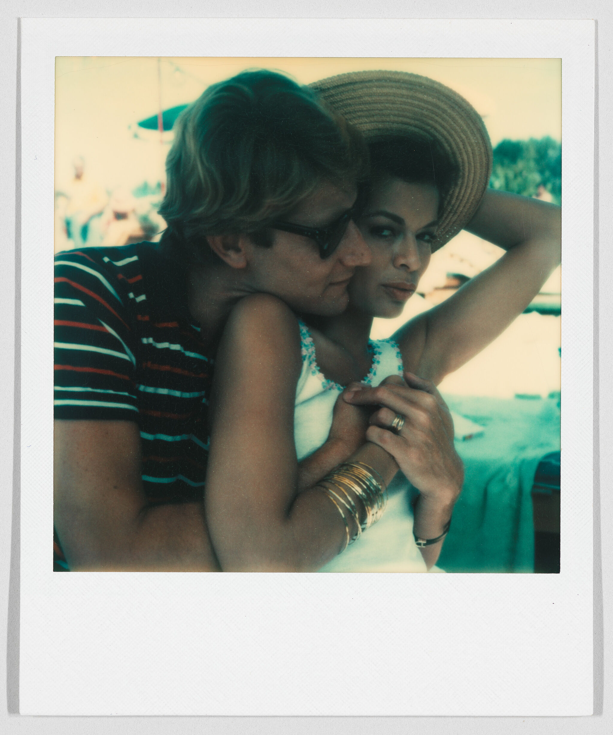 A polaroid image shows a man embracing a woman from behind, his neck leaning on her shoulder. He wears sunglasses and she holds a straw hat to her head, together with bright light and turquoise behind them, gives the impression of a summer day. 