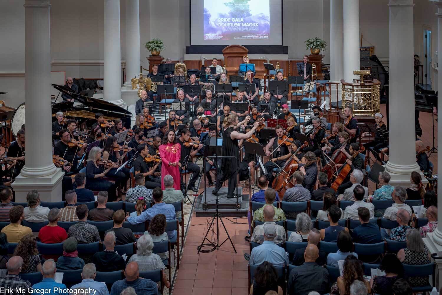 The Queer Urban Orchestra performs in a church-like concert hall filled with seated audience members. At the center, a conductor leads the ensemble while a vocalist in a bright pink outfit stands near the front of the stage. Musicians playing strings, brass, percussion, and piano are arranged across the space beneath tall white columns and a projected screen reading “Pride Gala Double… Magick.”  