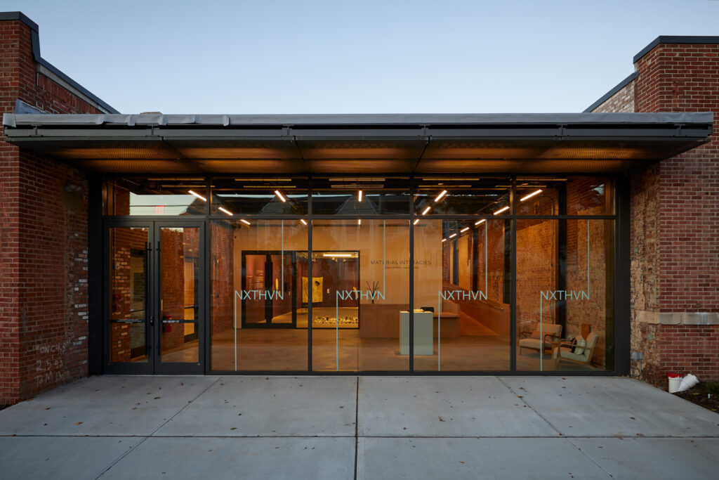Modern glass-front art gallery with exposed brick walls and warm interior lighting at dusk.
