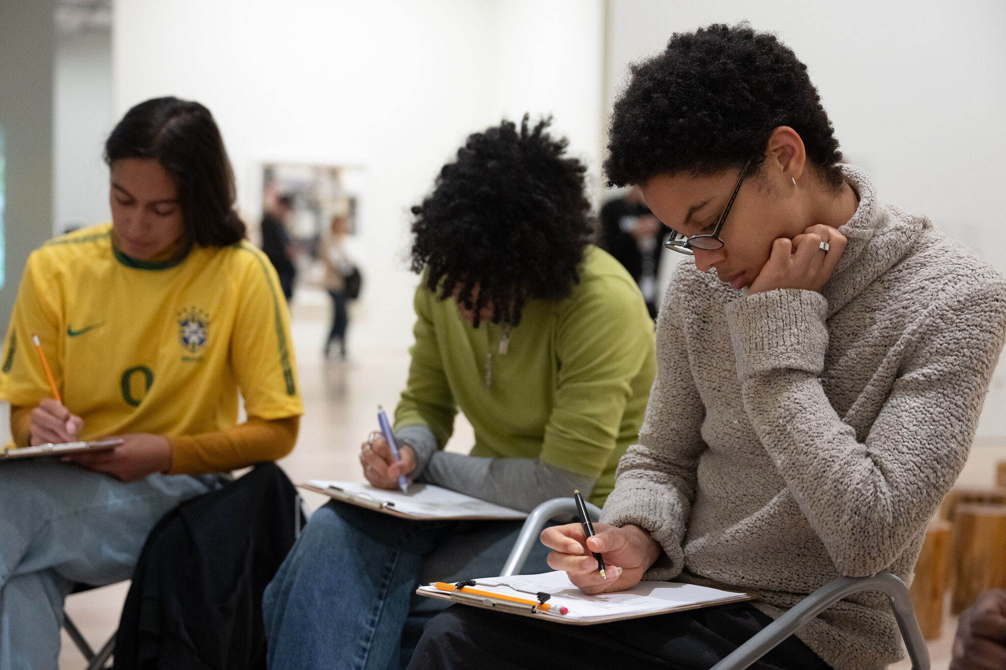 Three students sit closely and concentrate while drawing or writing on clipboards.