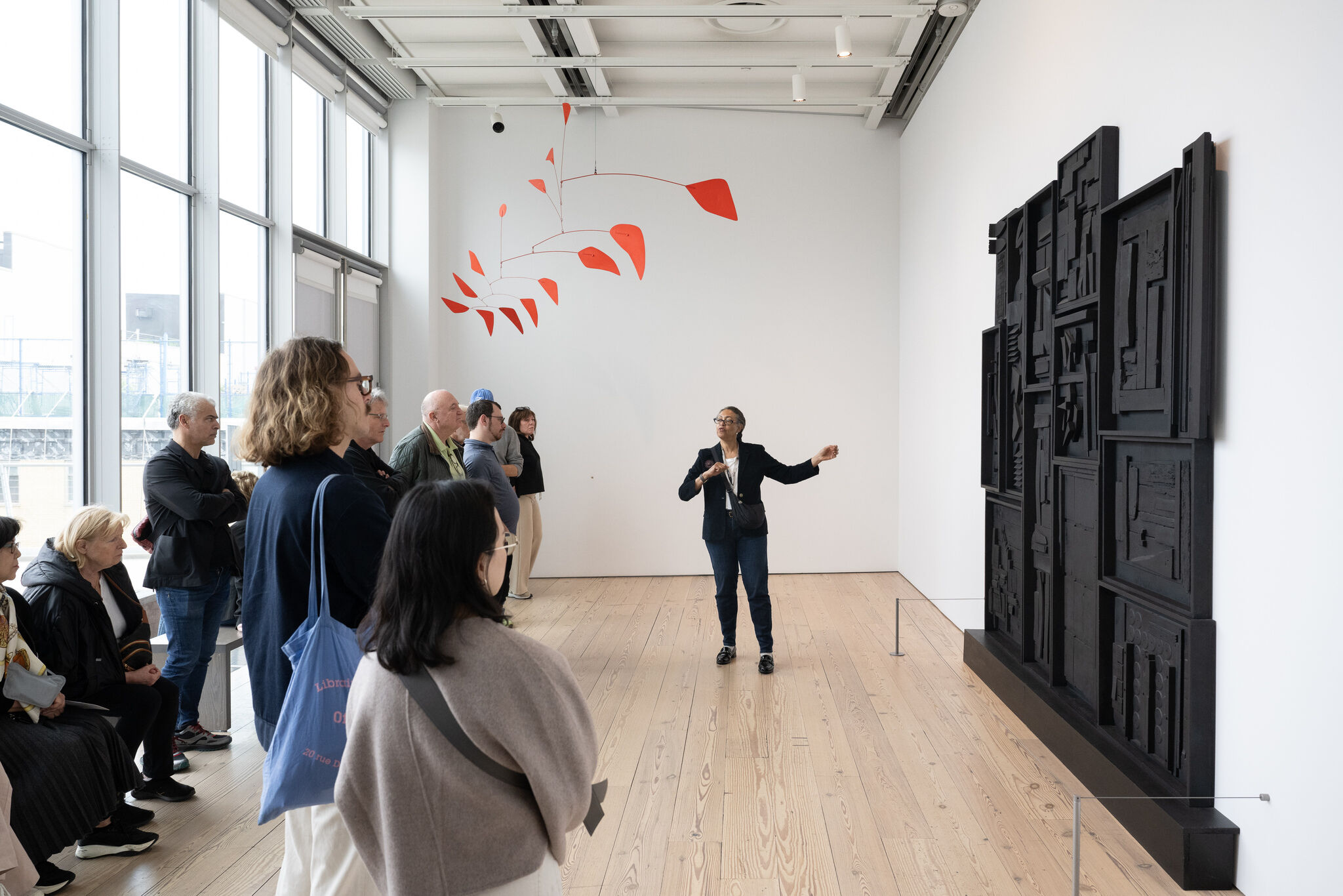 A museum guide gestures toward a red hanging mobile and black relief while visitors listen.