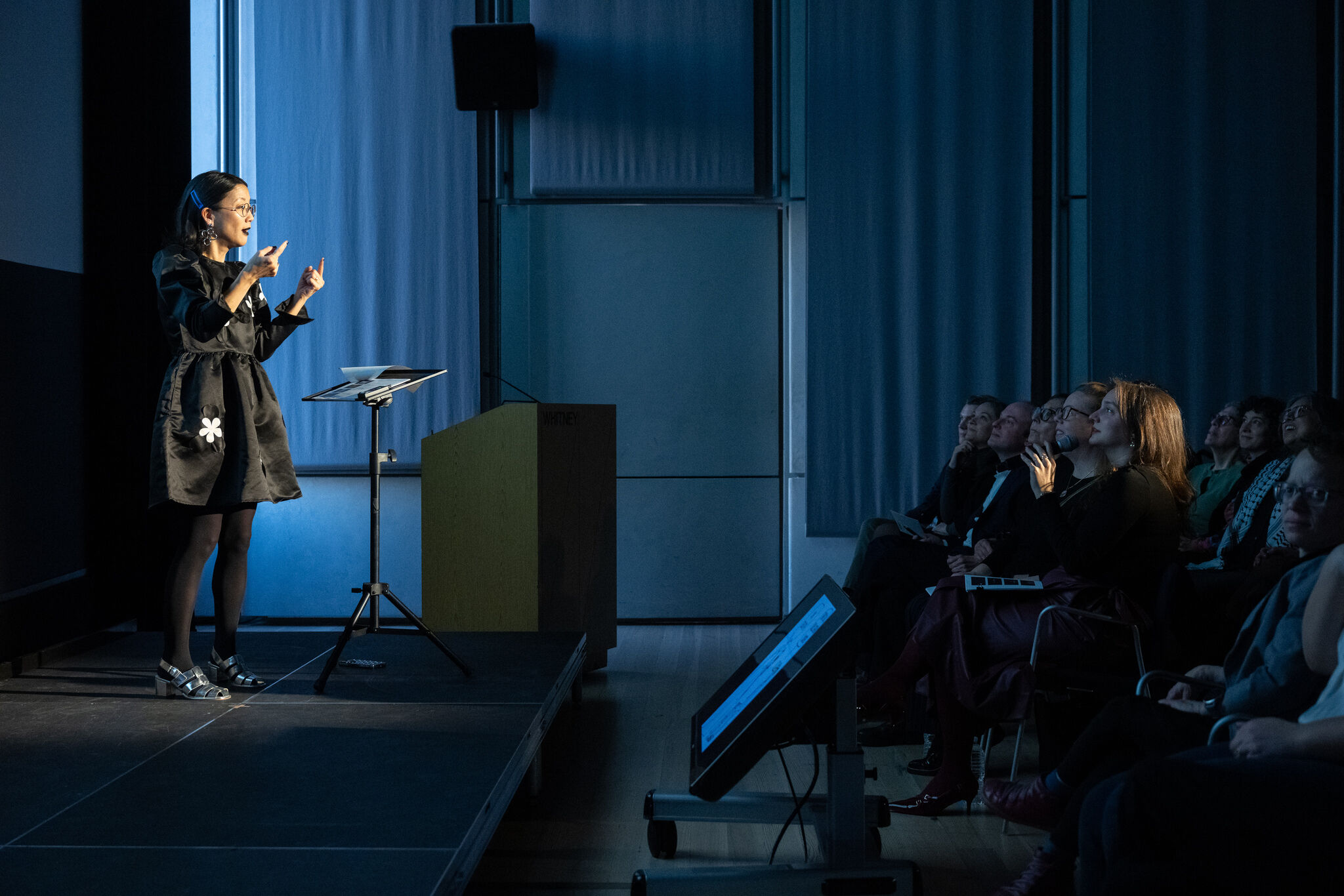 Woman speaker gestures passionately on stage while audience watches attentively under blue lighting.