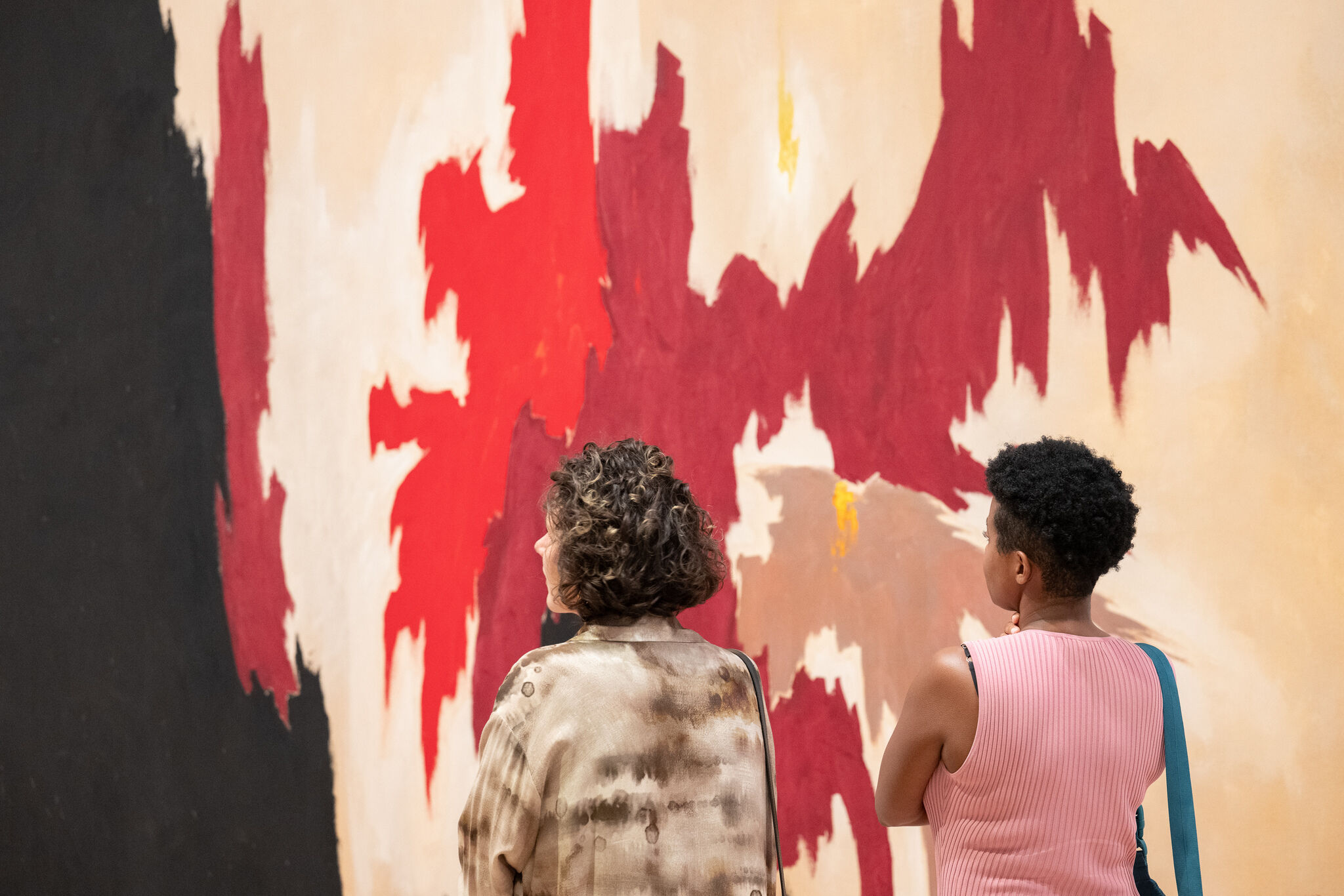 Two people stand facing a large abstract red and beige painting in a gallery.