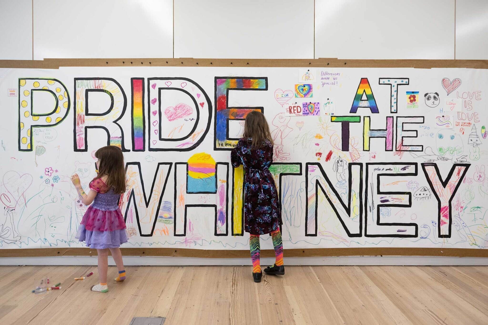 Two children color a large wall mural reading Pride at the museum with crayons.