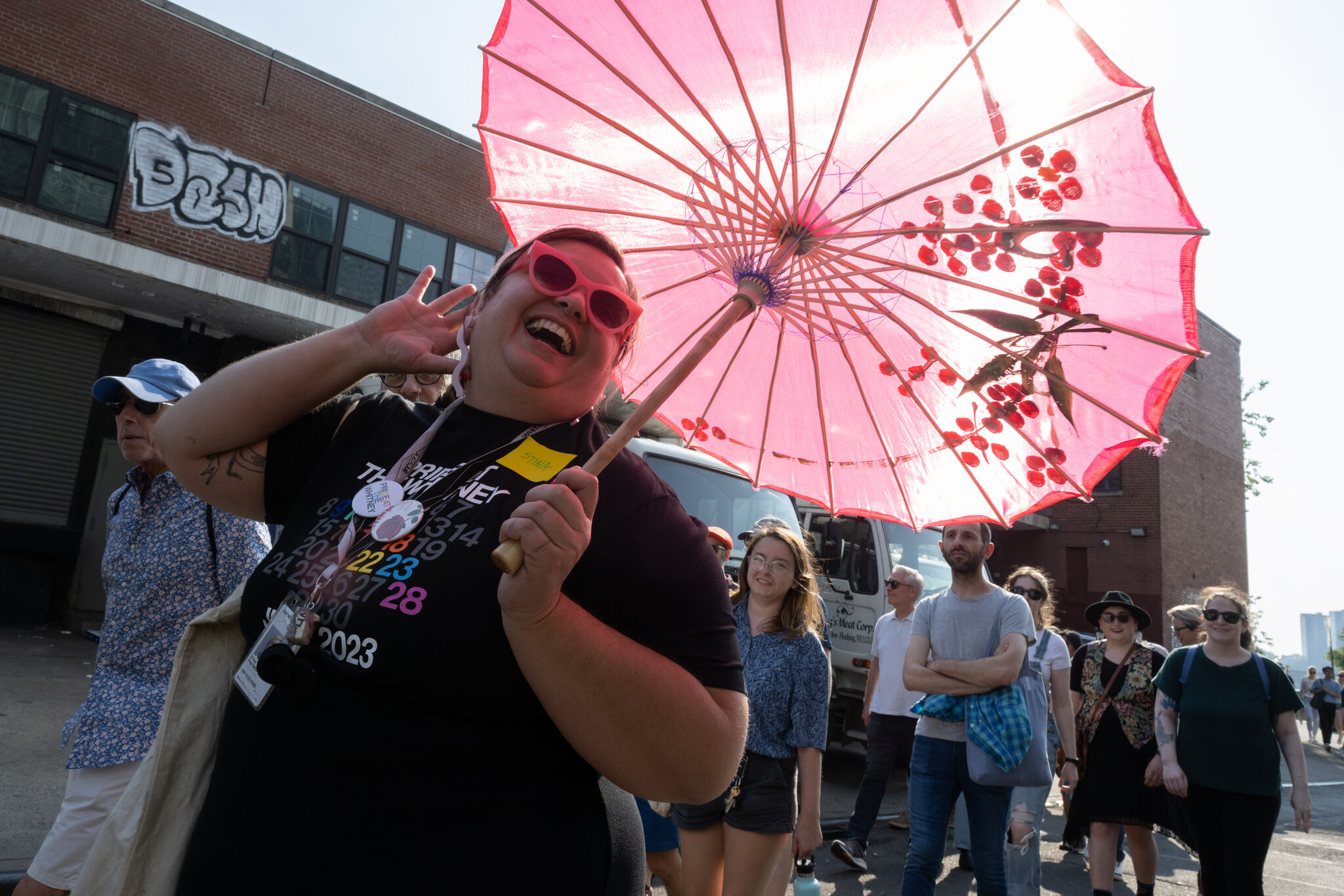 Person wearing pink sunglasses laughs while holding a decorative pink umbrella during a street march.
