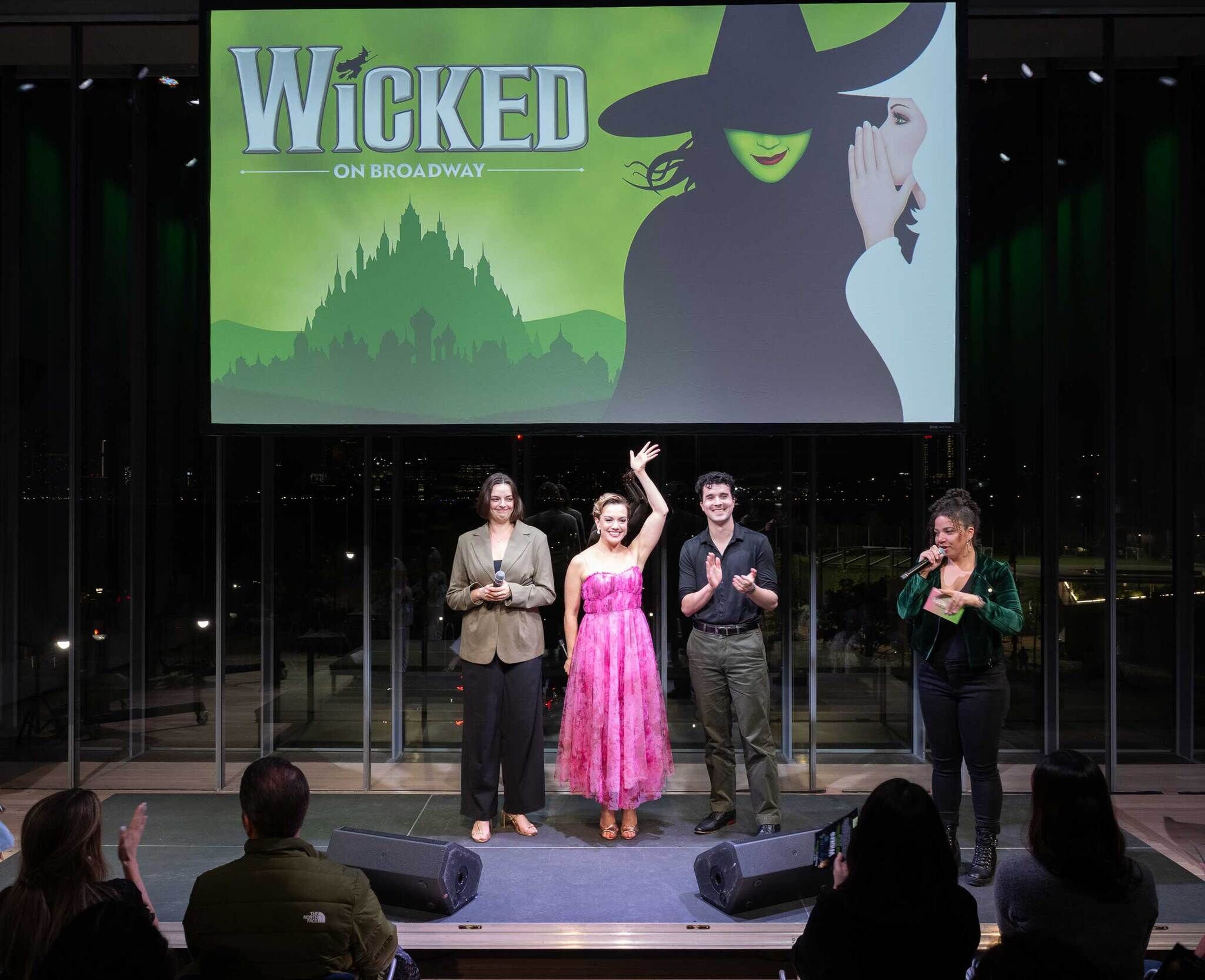 Performers from WICKED on Broadway stand onstage at the Whitney Museum during a live performance event, with a large WICKED projection behind them and audience members seated in front of the stage. 