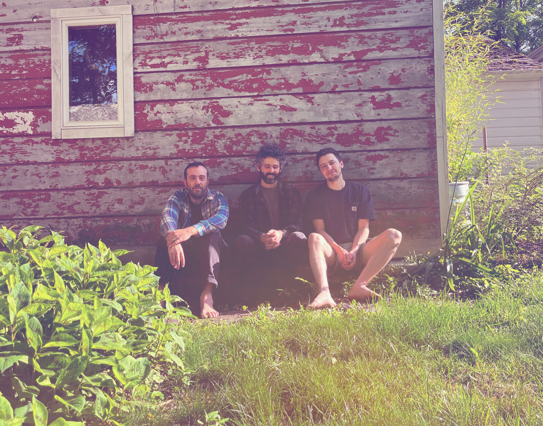 The trio behind Cowboy Sadness, Peter Silberman, Nicholas Principe, and David Moore, sit side by side, leaning on the exterior wall of a house and looking into the camera. 