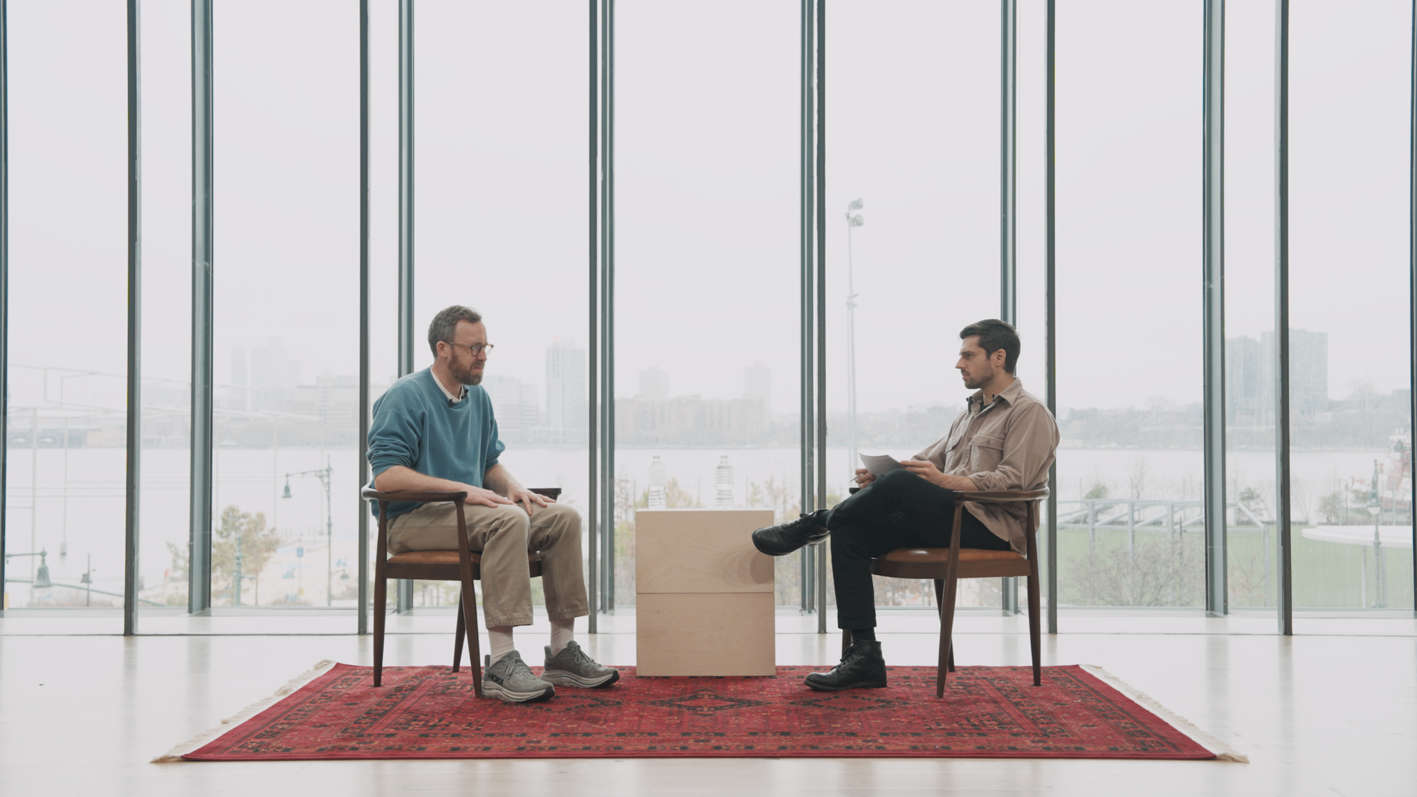 Two men sit facing each other in chairs by a window while one holds papers and they talk.