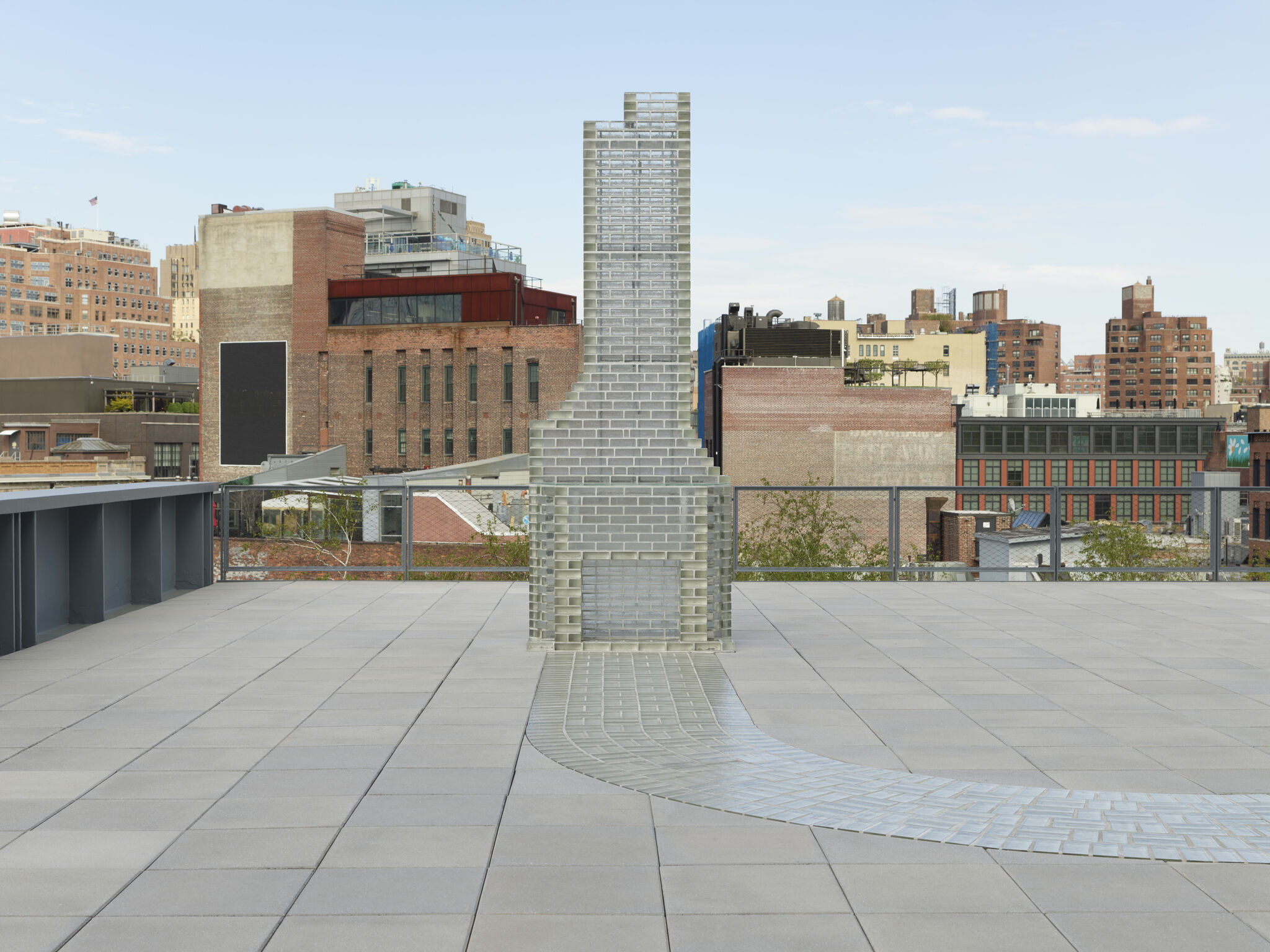 Installation view of Kelly Akashi's Monument (Altadena) (2026) at the Whitney Biennial 2026. A translucent glass sculpture resembling a stepped chimney or architectural tower stands on the Whitney Museum's outdoor terrace. Made of stacked glass blocks, the structure rises from a broad base into a tall vertical form. A winding path of glass pavers extends from the foreground toward the sculpture, suggesting a walkway or stream. The work is set against a backdrop of low-rise buildings and the Manhattan skyline beneath a pale blue sky.  
