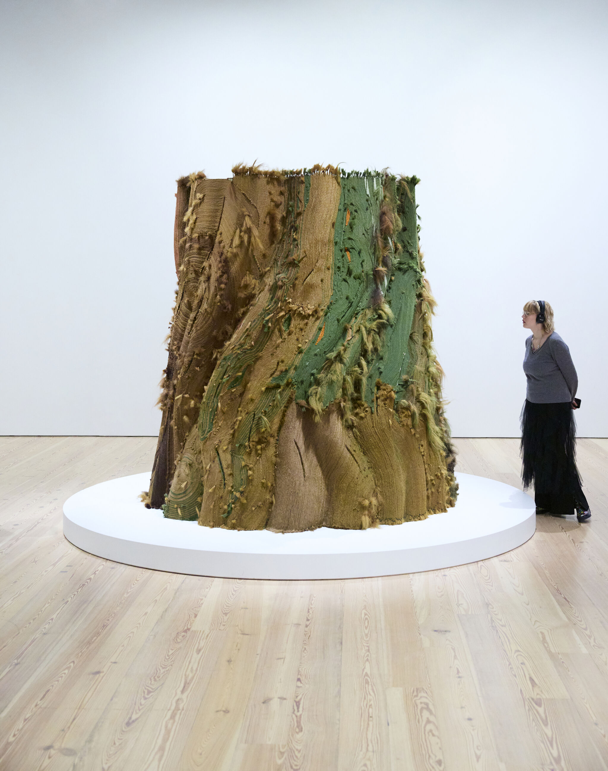 Person with headphones on gazes at a massive tree trunk sculpture made of brown and green woven hair.