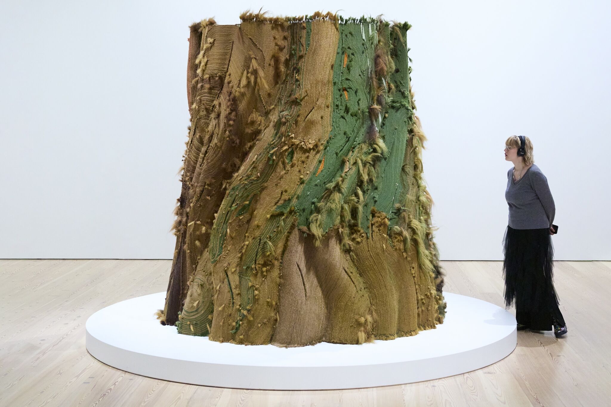 Person with headphones on gazes at a massive tree trunk sculpture made of brown and green woven hair.