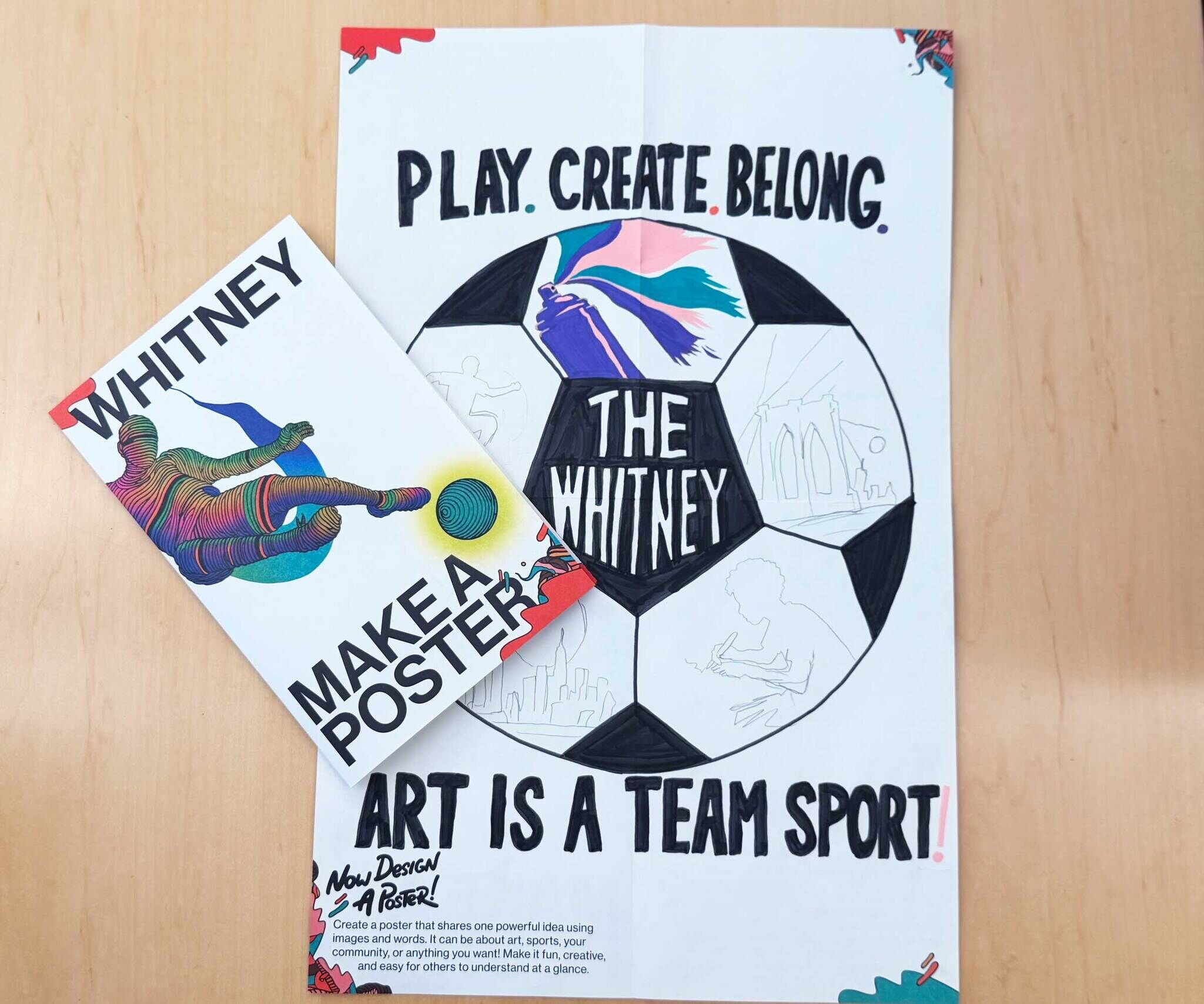 Two colorful posters sit on a table, one showing a soccer-ball design with the slogan 'Play. Create. Belong.'.