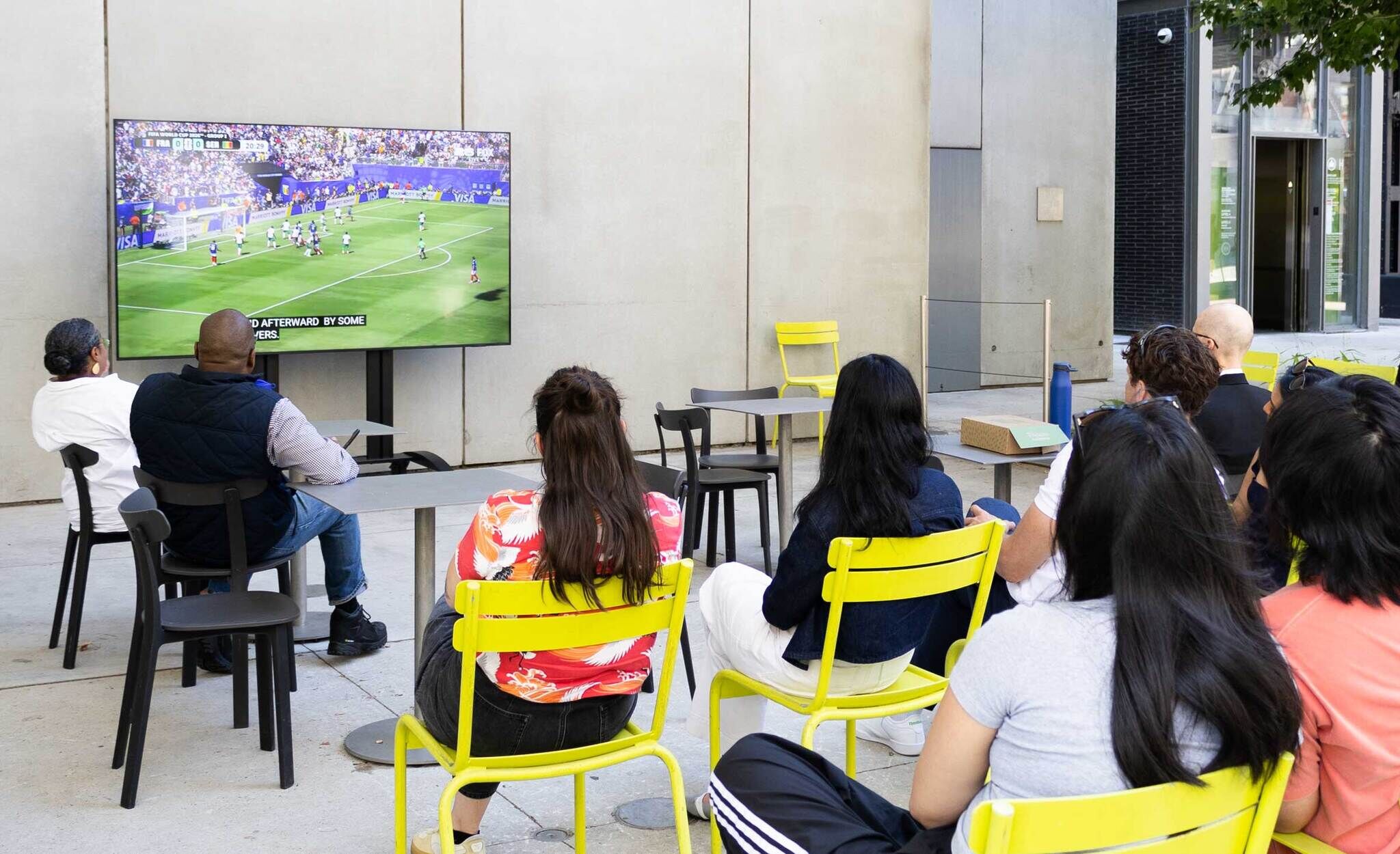 A group of people sit on yellow chairs watching a soccer match on a large outdoor screen.