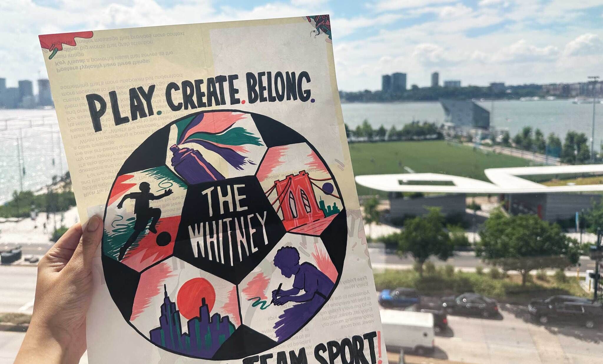 Hand holding colorful flyer reading "Play. Create. Belong." with waterfront and city skyline behind.