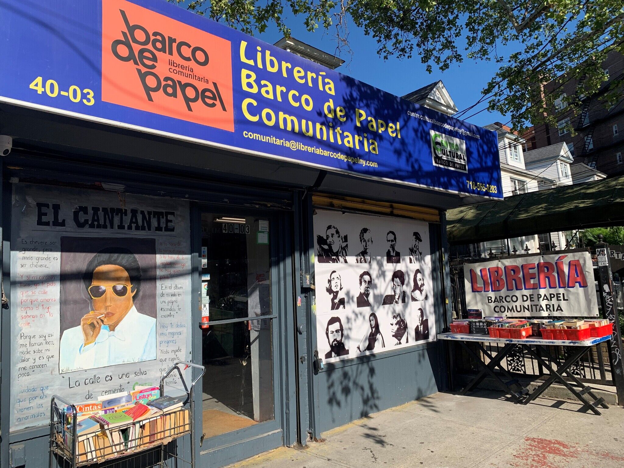 Photo of Librería Barco de Papel Comunitaria, a community bookstore with a blue and orange sign, a display table of books outside, and a storefront with black-and-white portraits of Spanish-speaking literary figures and a poster titled "El Cantante" with handwritten text.
