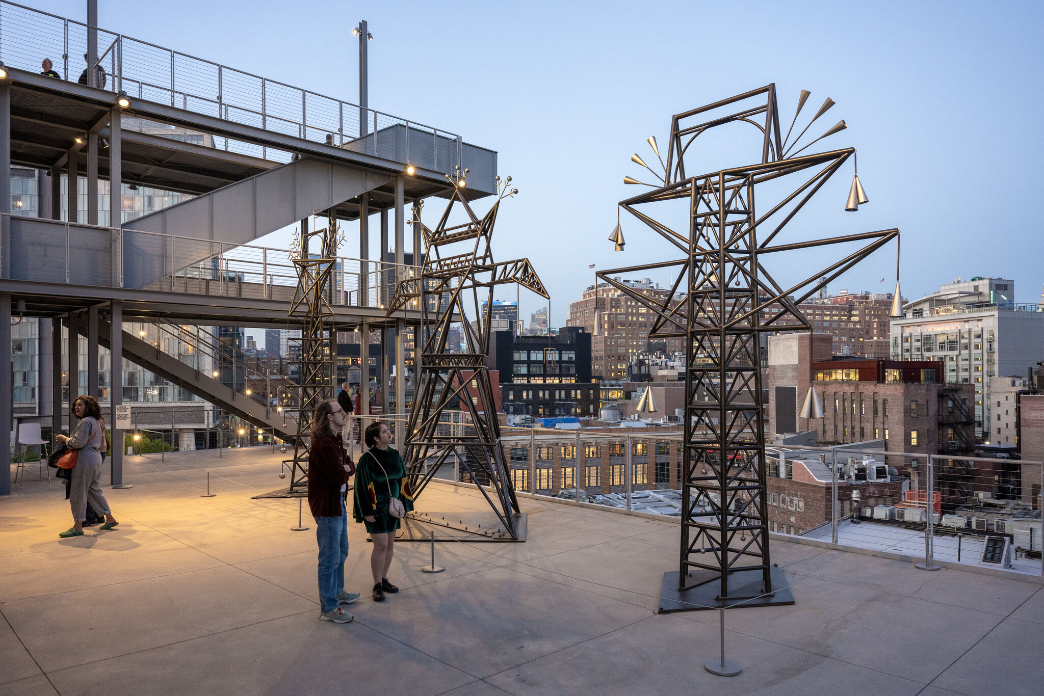 In the foreground, visitors look at transmission-tower sculptures on a terrace, with the west Manhattan cityscape in the background.