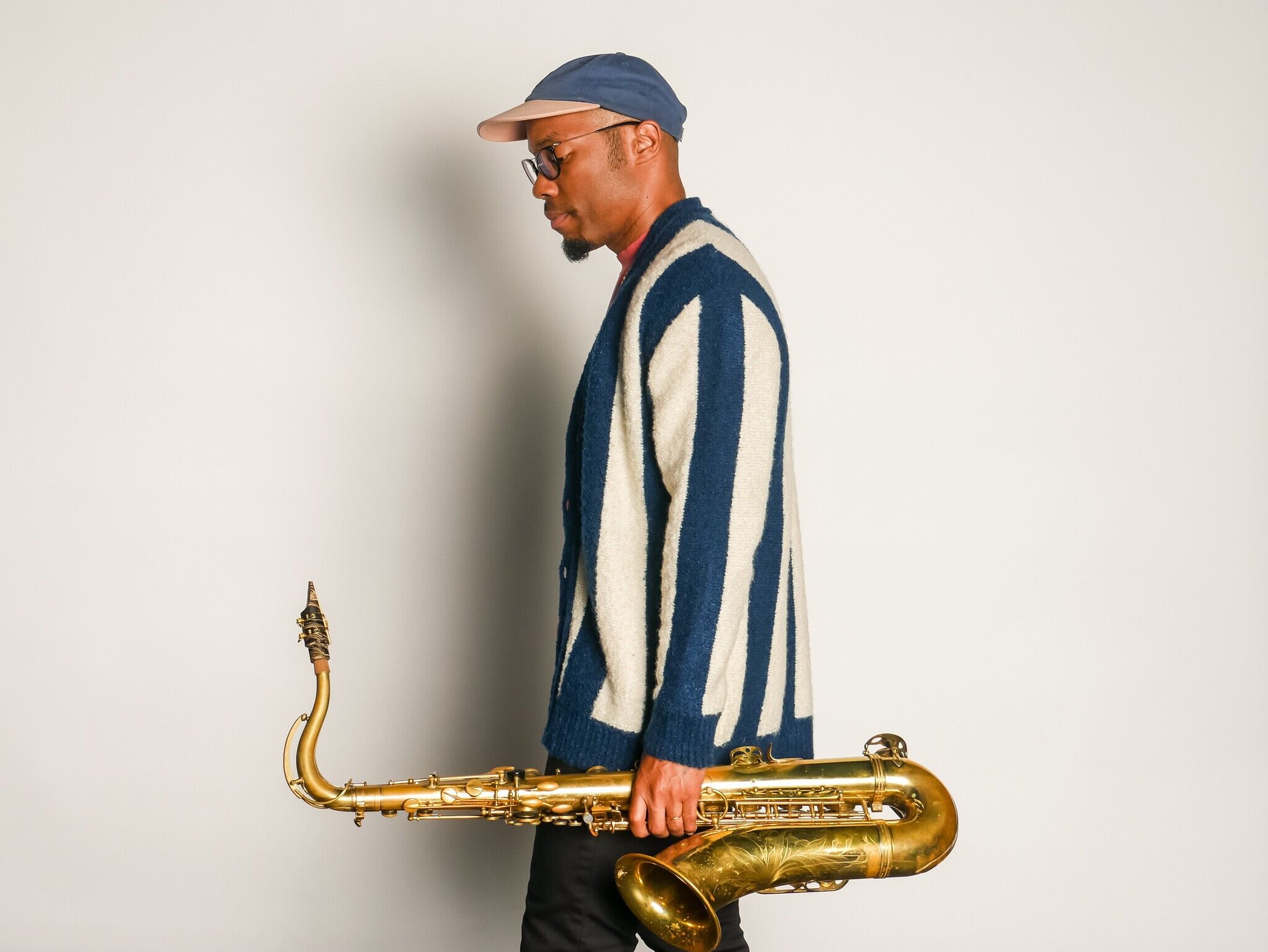 FKAjazz wearing a striped blue shirt and blue hat, looking to the side and carrying his saxophone