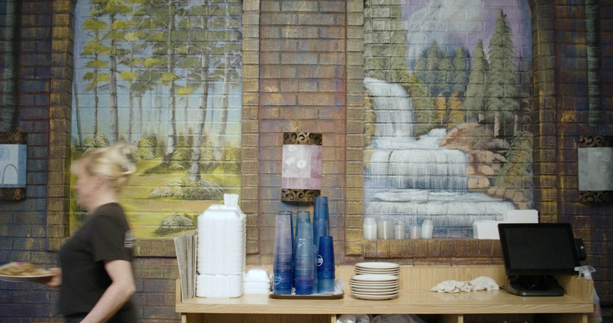 A server walks past a counter with stacked cups and plates in front of a mural on a brick wall of a tropical island and a lush waterfall.