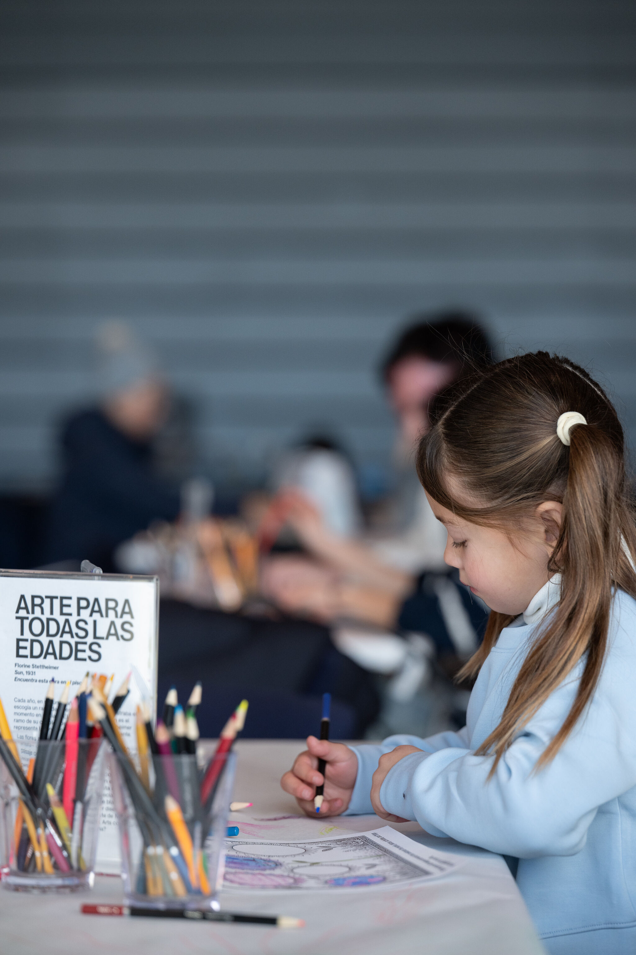 A young child sits at a table during the Whitney Museum's Free Second Sunday program, concentrating on a drawing with colored pencils. Containers of drawing materials and an all-ages artmaking sign are in the foreground, while other visitors creating art together appear softly blurred in the background. 