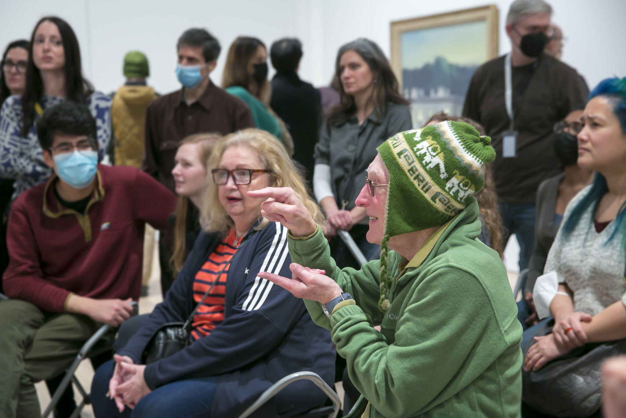 Elderly person in green hat gestures passionately while seated among a diverse group of masked and unmasked listeners.