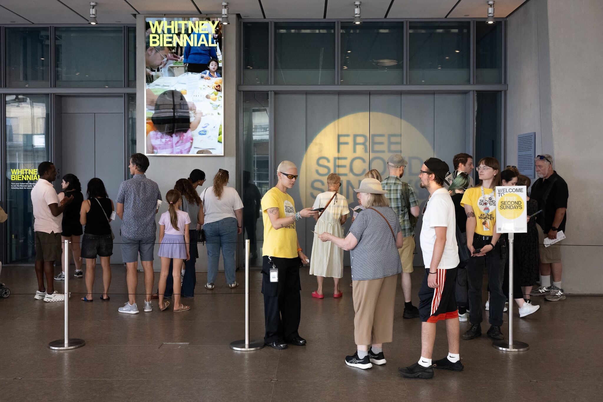 Visitors queue at the Whitney museum entrance while staff in yellow check tickets for admission.