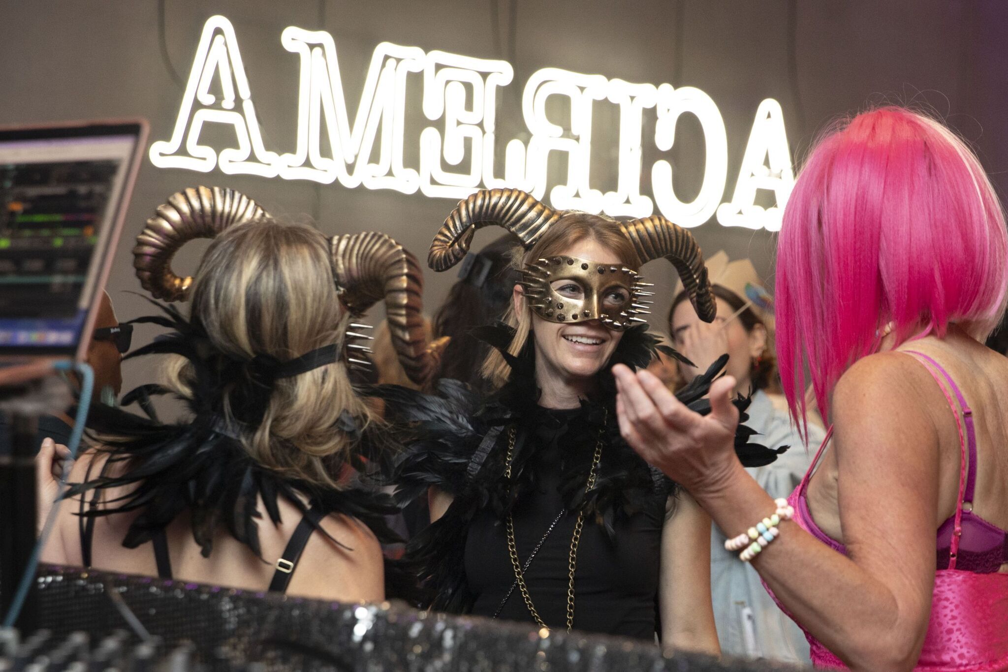 Two people in gold and black horns and masquerade masks speak happily with a person in a hot pink wig and top. 