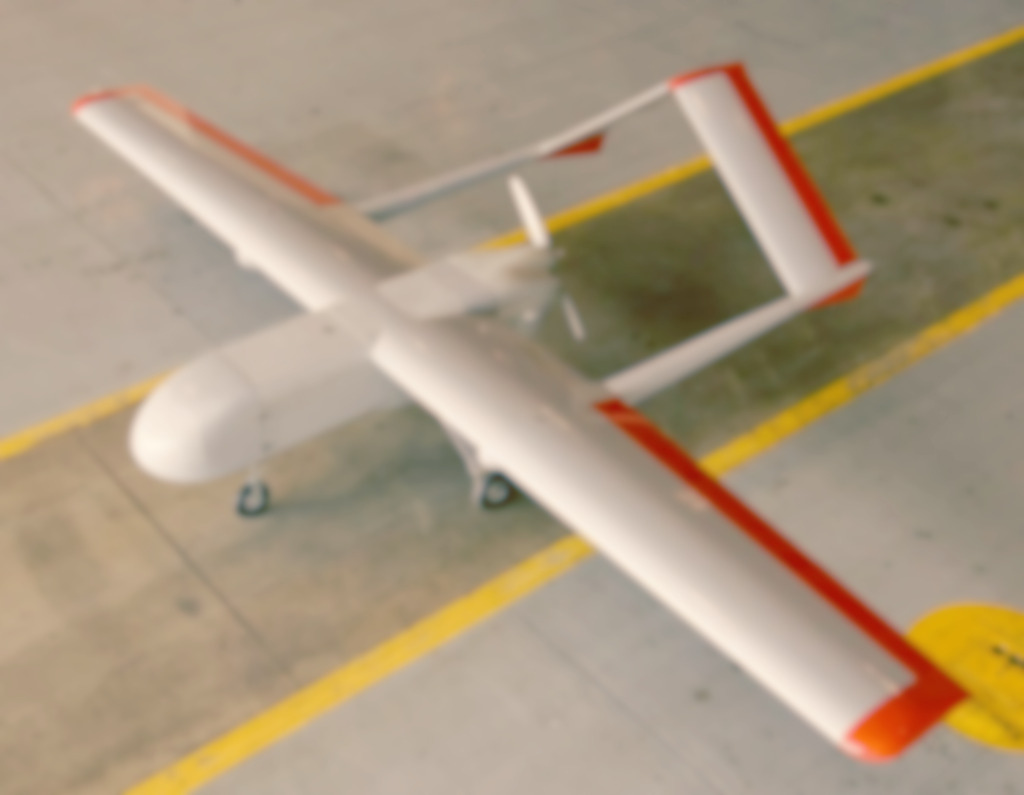 A white twin-boom unmanned aircraft with orange wingtips parked on a hangar floor.