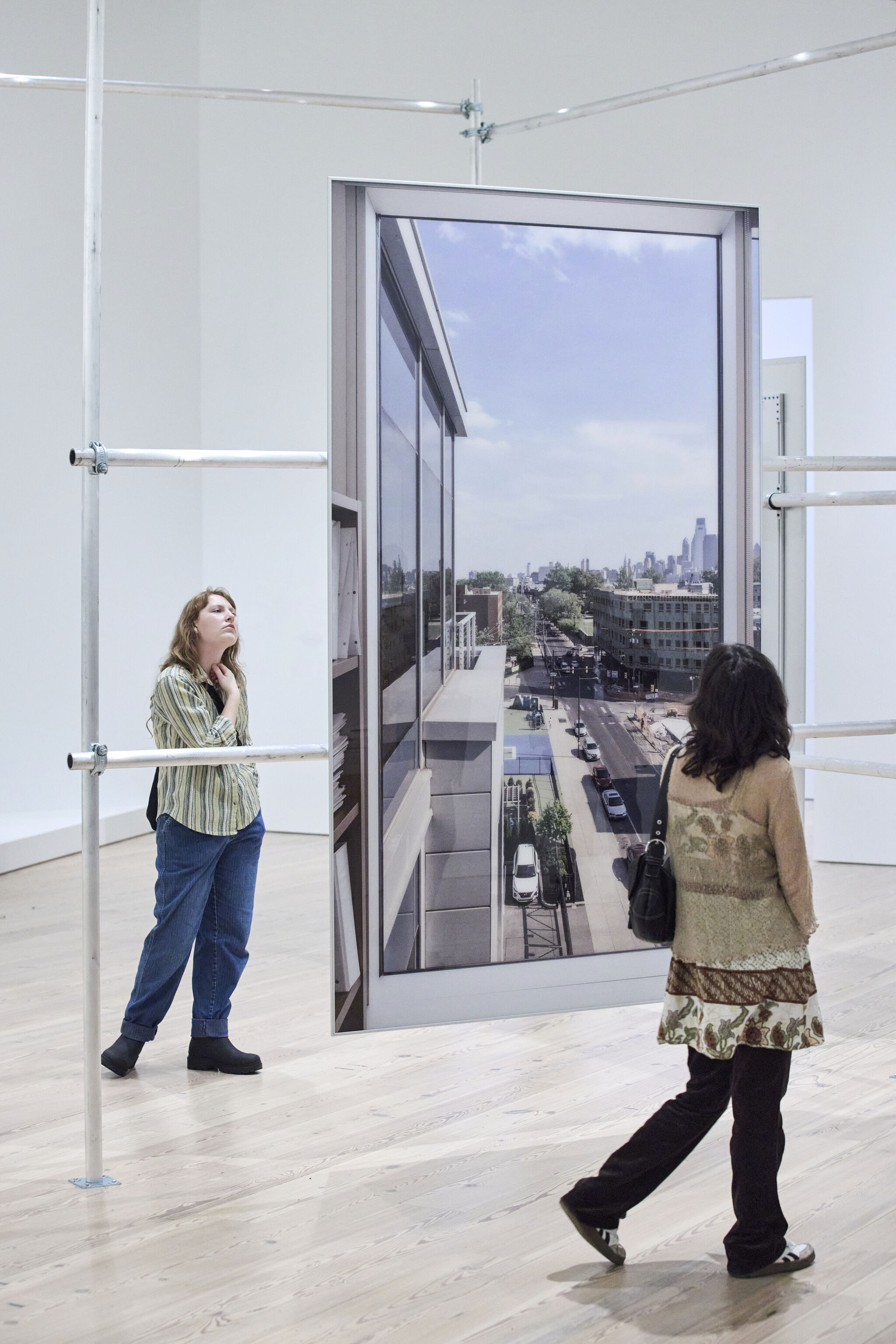 A large color photograph depicts the view from an office window looking out over a street in Philadelphia. The photograph is displayed in the middle of a white walled gallery on a silvery metal armature. Two female museum visitors look at the photograph from different perspectives.