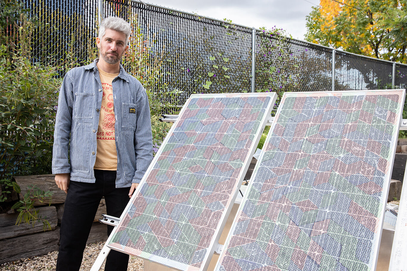 Alex Nathanson stands outdoors beside two large solar panels covered with colorful, hand-created geometric patterns. The panels are displayed on angled stands in front of a chain-link fence surrounded by plants and autumn foliage. 