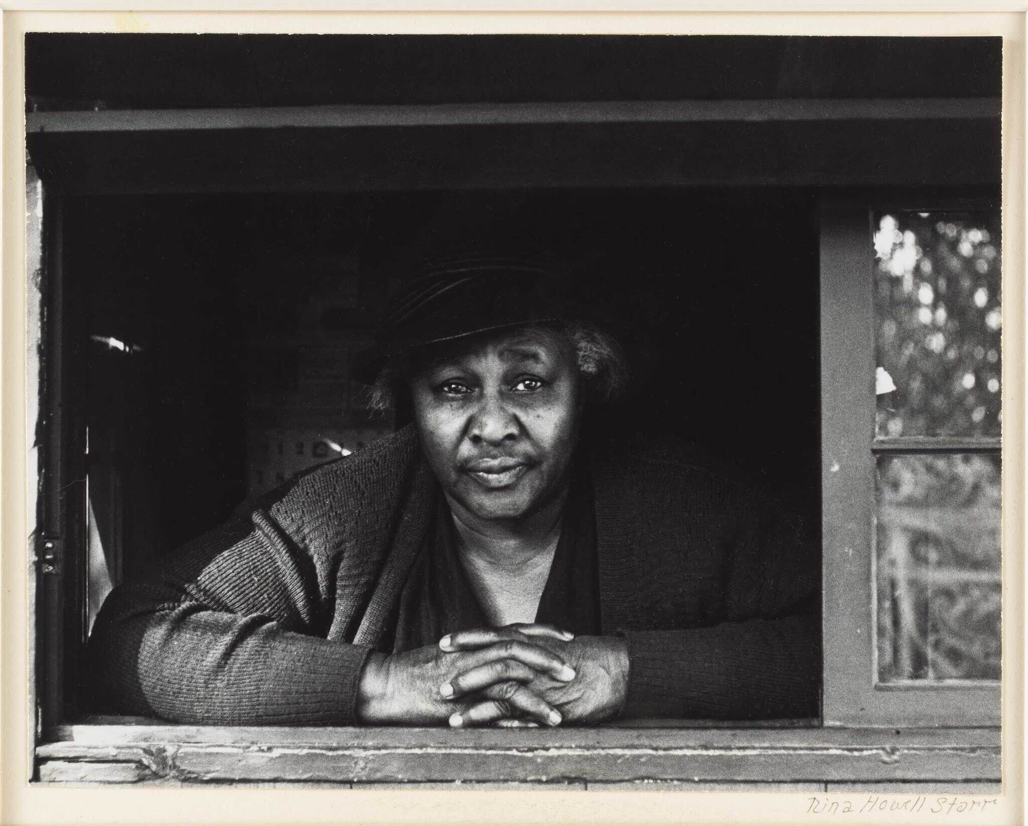 Older woman leaning on a windowsill with folded hands, looking directly at the camera.