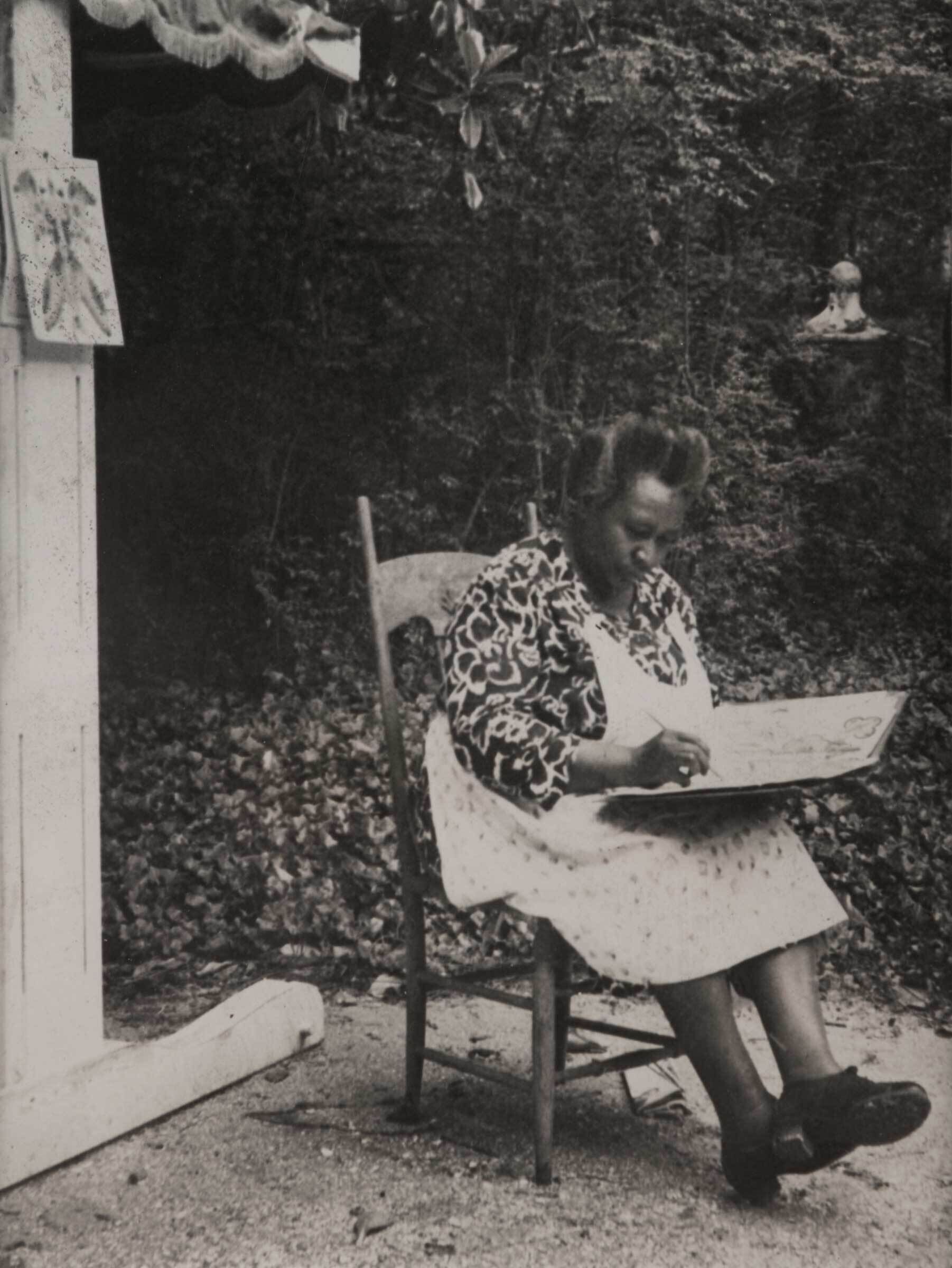 An older woman sits on a wooden chair outdoors, drawing on a large sketchpad.