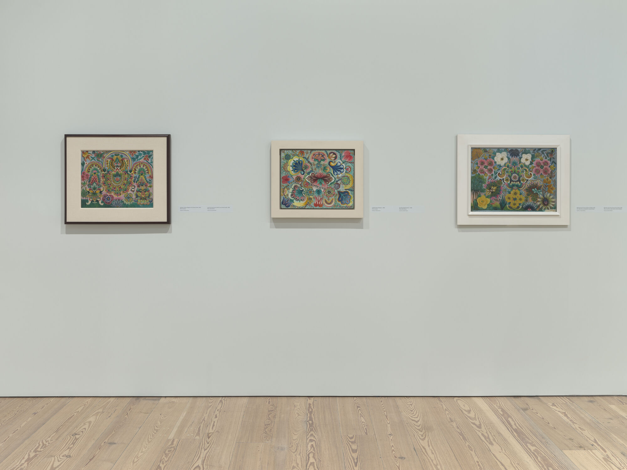Three small framed colorful floral paintings hang evenly spaced on a pale museum wall.