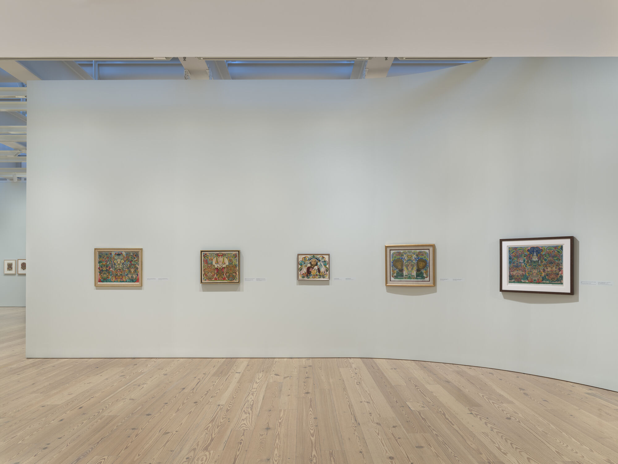 Five small colorful framed paintings hang evenly spaced on a white gallery wall above a wooden floor.