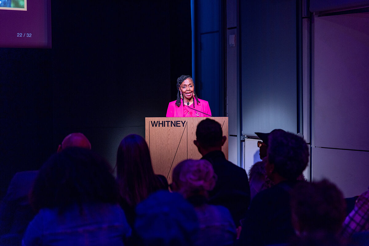 Donna Walker-Kuhne speaking from behind a podium, wearing a pink blazer and blouse and statement earrings. The podium is inscribed with "WHITNEY."