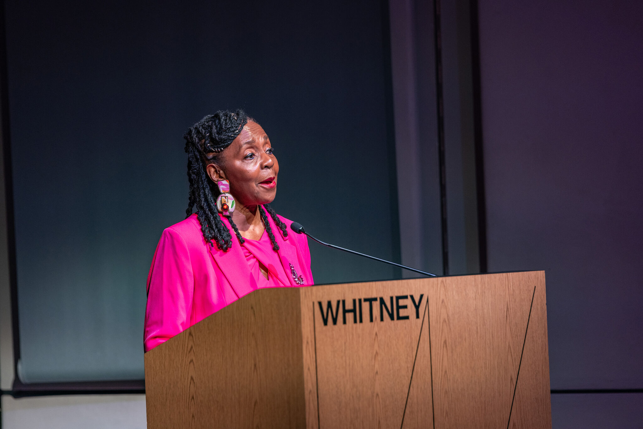 Donna Walker-Kuhne speaking from behind a podium, wearing a pink blazer and blouse and statement earrings. The podium is inscribed with "WHITNEY."