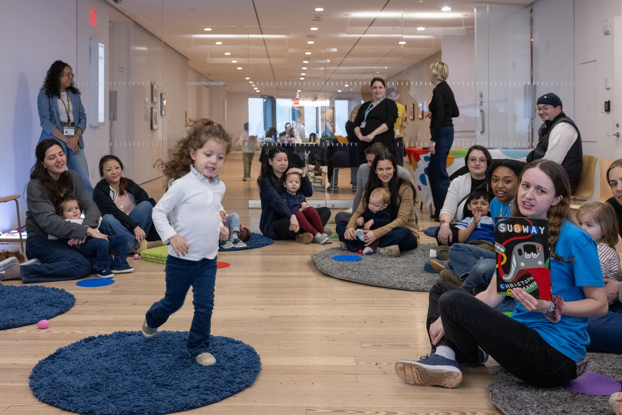 Families with young children gather on colorful rugs for Circle Time at the CMOM Pop Up Play Lounge at the Whitney. A child moves across a blue rug while an educator seated nearby holds up a picture book for the group. 
