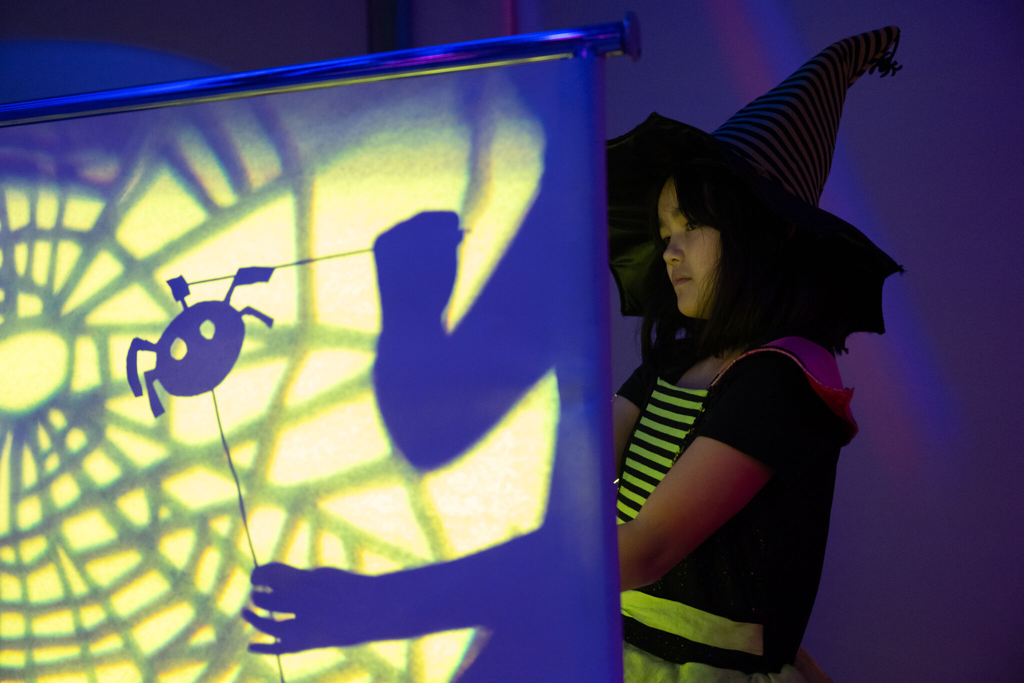 A child wearing a pointed witch hat creates a shadow puppet scene, casting the silhouette of a small creature and their own hands onto a brightly lit screen patterned with a spiderweb. 
