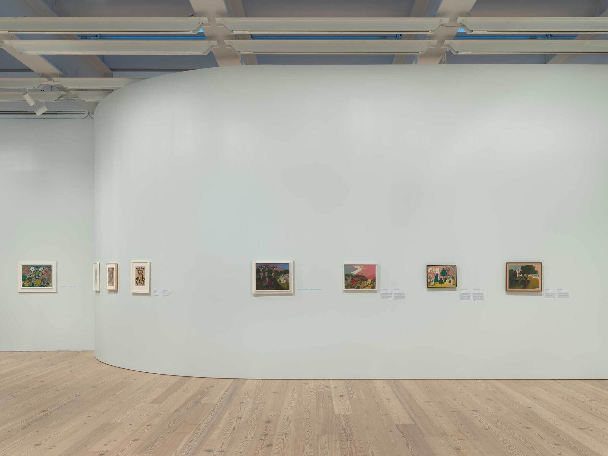 A curved white gallery wall displays a row of small framed paintings above a wooden floor.