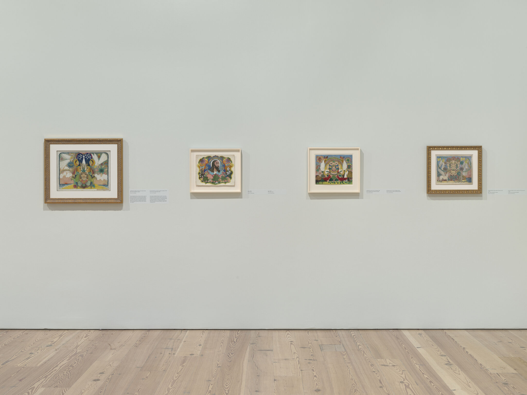 Four small colorful framed paintings hang evenly spaced on a pale gallery wall above a wooden floor.