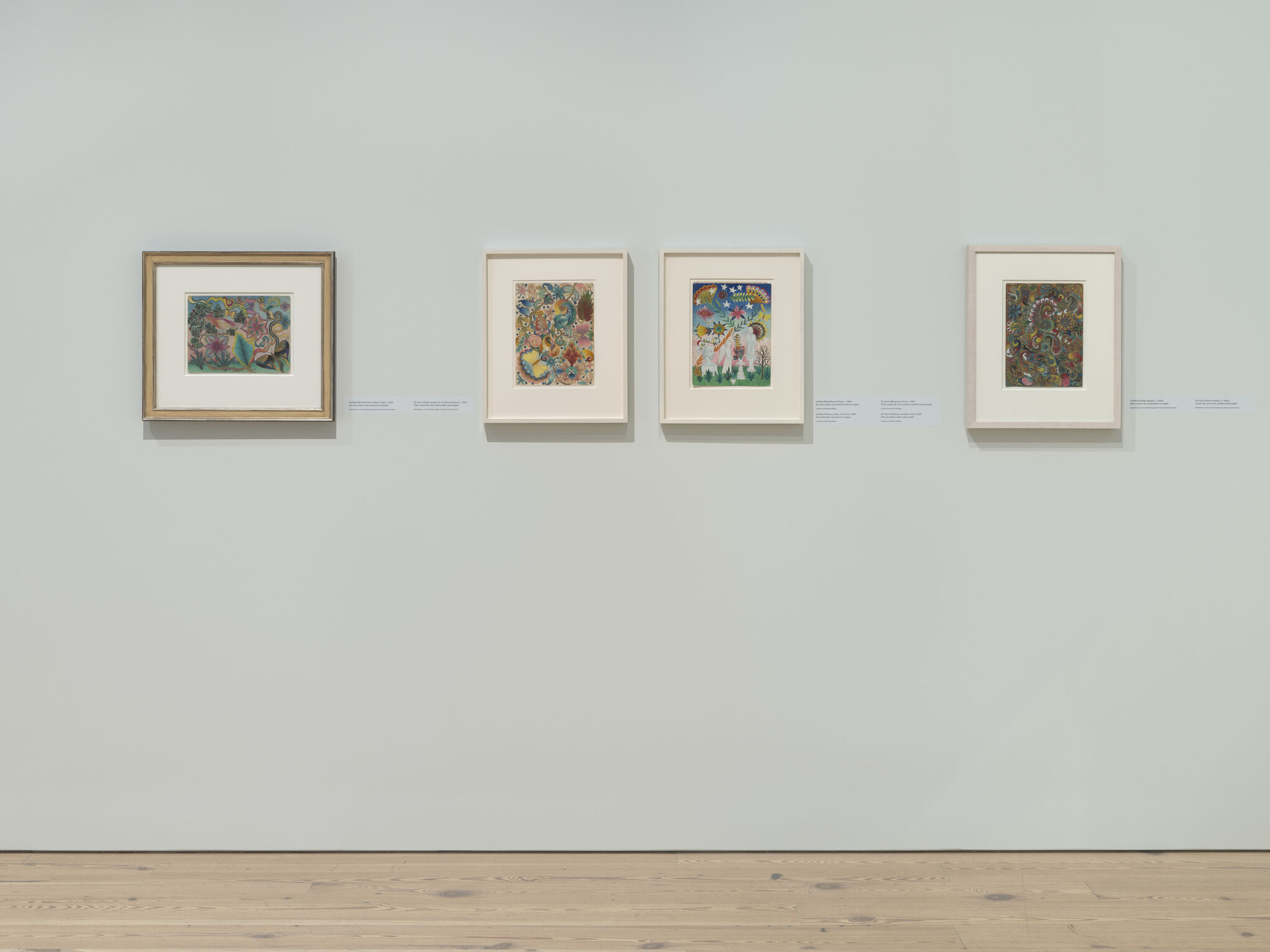 Four colorful framed artworks hang evenly on a pale gallery wall above a wooden floor.