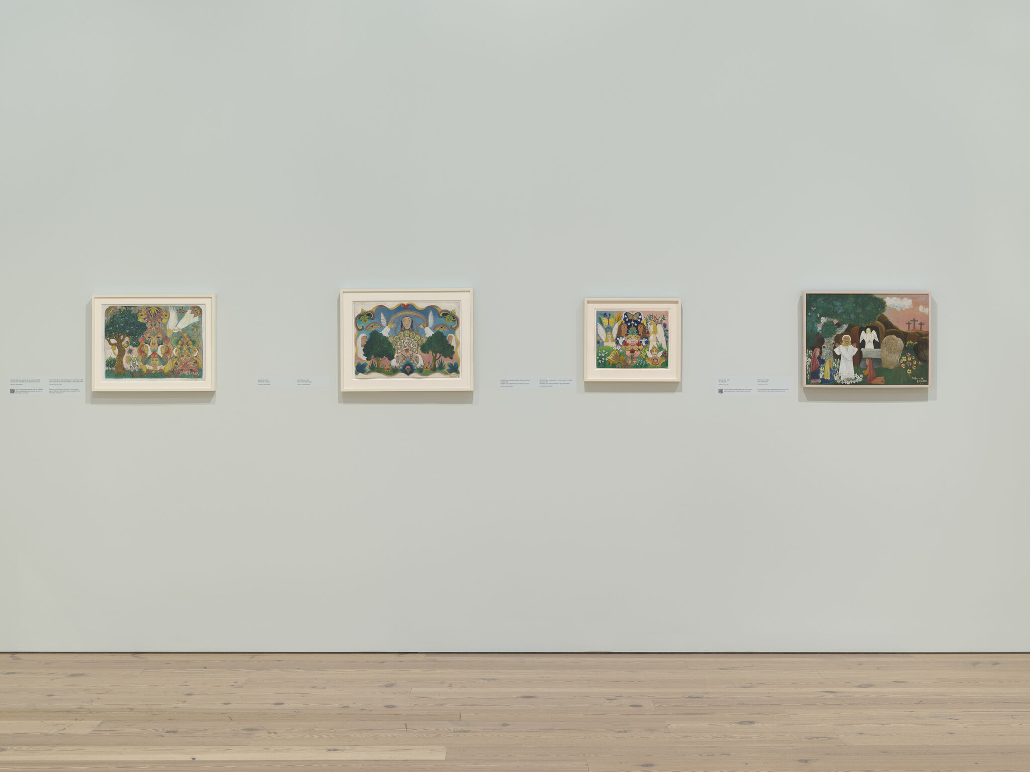 Four small colorful framed paintings hang evenly spaced on a pale gallery wall above wooden floor.