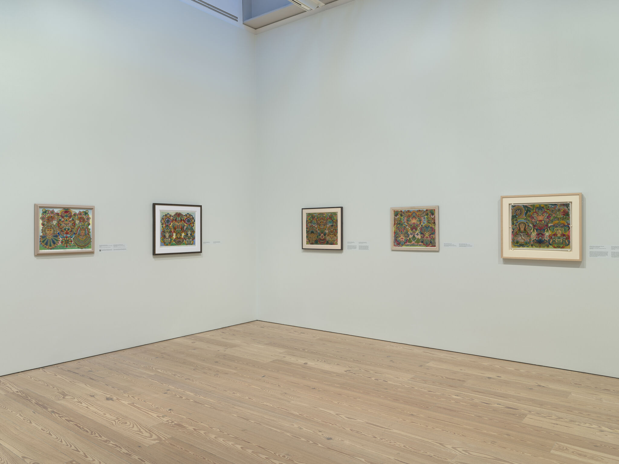A row of five framed colorful folk-art paintings displayed along a pale gallery wall.