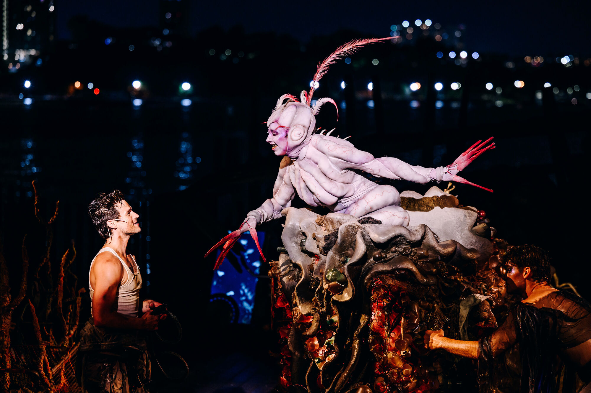 A pale, insect-like performer with long red claws reaches toward a shirtless man on stage.